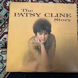 May include: A square vinyl record album cover titled The Patsy Cline Story. The cover features a portrait of Patsy Cline in a tan shirt against a gold background. The text reads The Patsy Cline Story and Deluxe 2-Record Set in black font.