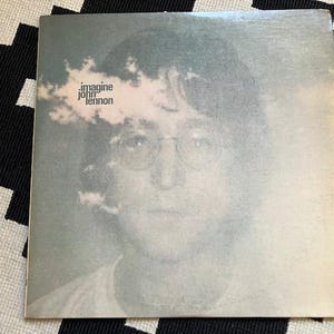 May include: Album cover for John Lennon's "Imagine." The cover features a faded, grayscale portrait of Lennon wearing glasses. The title is printed in a stylized font with a cloud-like design. The album is on a black and white patterned surface.