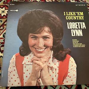 May include: A vintage vinyl record album cover featuring Loretta Lynn. The cover displays a portrait of the artist with dark hair, wearing a white shirt with red polka dots and red suspenders. Text includes I LIKE 'EM COUNTRY and song titles.