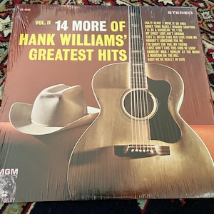 May include: A vinyl record album cover titled 14 More of Hank Williams' Greatest Hits Vol. II. The cover features a brown background, a white cowboy hat, and an acoustic guitar. The tracklist is printed in yellow text on the upper right side.