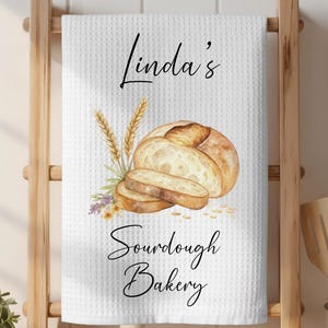 May include: White waffle-weave kitchen towel with a watercolor illustration of a loaf of bread and sliced bread. The towel has the text "Linda's Sourdough Bakery" in black script.