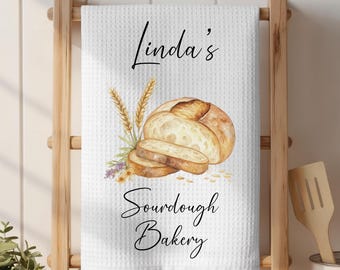 May include: White waffle-weave kitchen towel with a watercolor illustration of a loaf of bread and sliced bread. The towel has the text "Linda's Sourdough Bakery" in black script.