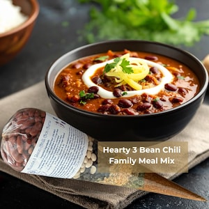 May include: A clear plastic cone-shaped package containing dried beans and spices labelled Hearty 3 Bean Chili Family Meal Mix, positioned next to a black bowl filled with chilli topped with a swirl of sour cream, shredded cheese, and coriander.