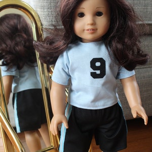 Pode incluir: Uma boneca com cabelo castanho comprido vestindo uma camiseta azul claro com um n&uacute;mero 9 preto na frente e shorts pretos com uma faixa azul claro na lateral.