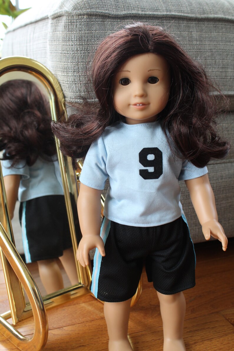Pode incluir: Uma boneca com cabelo castanho comprido vestindo uma camiseta azul claro com um n&uacute;mero 9 preto na frente e shorts pretos com uma faixa azul claro na lateral.