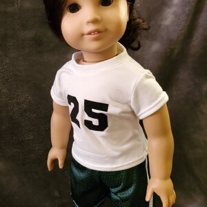 Pode incluir: Uma boneca vestindo uma camisa branca com o n&uacute;mero 25 na frente e shorts esportivos verdes com uma faixa branca na lateral.