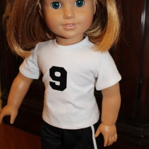 Pode incluir: Uma boneca vestindo uma camiseta branca com um n&uacute;mero 9 preto na frente e shorts esportivos pretos com uma faixa branca na lateral.