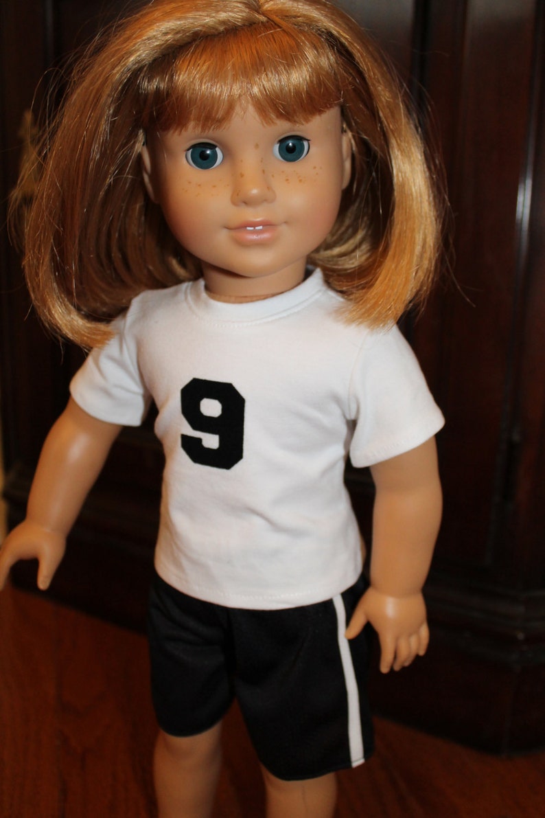 Pode incluir: Uma boneca vestindo uma camiseta branca com um n&uacute;mero 9 preto na frente e shorts esportivos pretos com uma faixa branca na lateral.