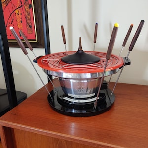 May include: A vintage red and black fondue pot with a silver bowl and eight wooden fondue forks. The pot is on a wooden table.
