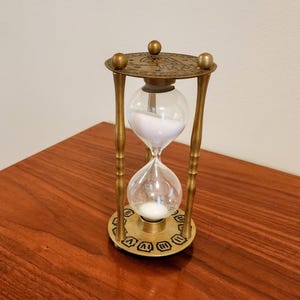 May include: An hourglass with a brass-toned metal frame featuring three vertical pillars and circular top and bottom plates. The top plate displays engraved zodiac symbols, while the bottom plate features Roman numerals. Clear glass bulbs contain white sand.