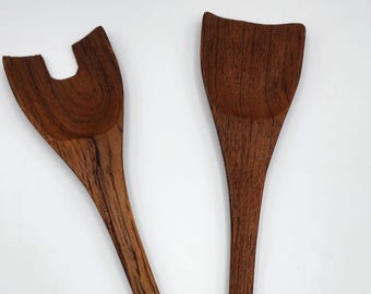 May include: A pair of dark brown wooden salad servers featuring a distinct grain pattern. One server has a wide, solid, curved head, while the other server features a notched, fork-like design with a central cutout, both with long, slender handles.