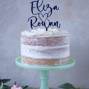 Pode incluir: Um bolo branco com cobertura de manteiga e um topo de bolo de acrílico preto que diz "Eliza Rowan" com um coração no meio.