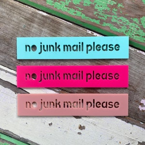 May include: Three rectangular signs in shades of blue, pink, and peach, each with the words "no junk mail please" cut out. The signs are arranged horizontally against a weathered wooden surface.