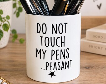 May include: A white cylindrical ceramic pen holder featuring the black printed text DO NOT TOUCH MY PENS ...PEASANT with a small star icon at the bottom. The holder is filled with several black retractable ballpoint pens with silver clips.