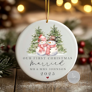 Puede incluir: Adorno navideño redondo de cerámica blanca. Presenta dos muñecos de nieve con sombreros y bufandas, frente a abetos. El texto dice "Our First Christmas Married", un nombre y el año 2025. Un pequeño corazón rojo está en la parte inferior.