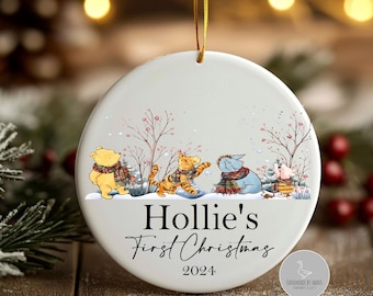 Könnte beinhalten: Runder weißer Weihnachtsbaumschmuck mit goldenem Faden. Er zeigt Winnie-the-Pooh-Figuren in einer Schneelandschaft mit dem Text "Hollie's First Christmas 2024". Die Figuren tragen Schals.