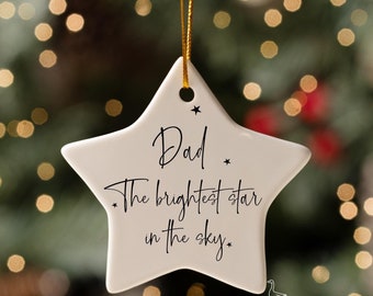 Puede incluir: Adorno navideño blanco en forma de estrella con el texto "Dad. The brightest star in the sky." en escritura negra. El adorno cuelga de un hilo dorado sobre un fondo borroso de árbol de Navidad.