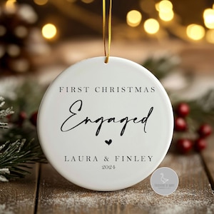 Puede incluir: Un adorno redondo de cerámica blanca que cuelga de una cinta dorada. El texto FIRST CHRISTMAS Engaged con un pequeño corazón, seguido de los nombres LAURA & FINLEY y el año 2024, está impreso en tinta negra en la parte frontal.