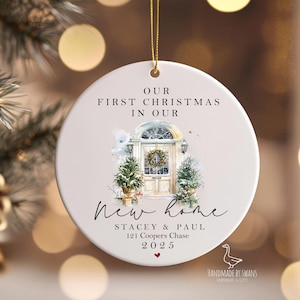 Puede incluir: Adorno navideño redondo y blanco con una cuerda dorada. El adorno presenta una ilustración en acuarela de una puerta principal con árboles de Navidad y el texto "Our First Christmas in Our New Home". Incluye nombres, dirección y el año 2025.