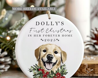 May include: A white ceramic Christmas ornament with a smiling golden retriever illustration. The ornament reads "Dolly's First Christmas in Her Forever Home 2025." It is adorned with a green and red holly wreath and a jute string for hanging.