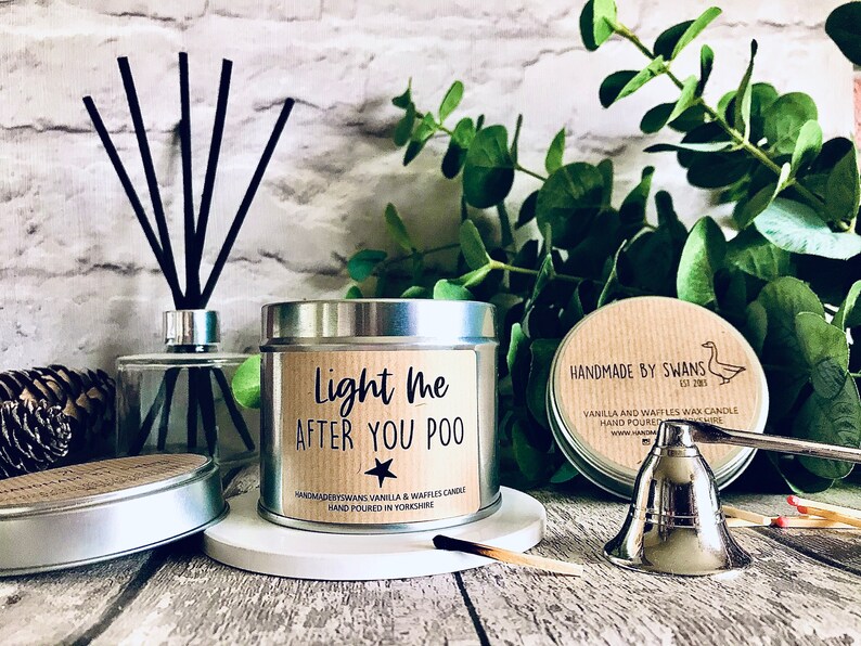 May include: A silver tin candle with a brown label that reads "Light me after you poo." The candle is surrounded by matches, a silver candle snuffer, and a diffuser.