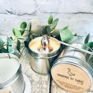 May include: Two silver tin candle jars with lids. One jar has a brown label with the text "HANDMADE BY SWANS EST 2013 VANILLA AND WAFFLES WAX CANDLE HAND POURED IN YORKSHIRE WWW.HANDMADEBYSWANS.COM". The other jar has a white label with the text "Christmas Home" and a small image of a Christmas tree.