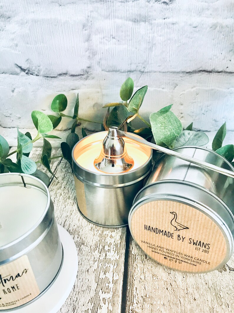 May include: Two silver tin candle jars with lids. One jar has a brown label with the text "HANDMADE BY SWANS EST 2013 VANILLA AND WAFFLES WAX CANDLE HAND POURED IN YORKSHIRE WWW.HANDMADEBYSWANS.COM". The other jar has a white label with the text "Christmas Home" and a small image of a Christmas tree.