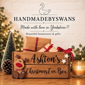 Puede incluir: Una caja de madera rectangular con el texto Ashton's Christmas Eve Box impreso en negro. Un recorte de reno de madera con una nariz roja est&aacute; sujeto al lado izquierdo. Dos paquetes de papel marr&oacute;n con cinta roja est&aacute;n dentro de la caja.