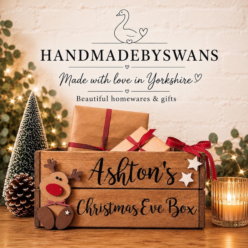 Puede incluir: Una caja de madera rectangular con el texto Ashton's Christmas Eve Box impreso en negro. Un recorte de reno de madera con una nariz roja est&aacute; sujeto al lado izquierdo. Dos paquetes de papel marr&oacute;n con cinta roja est&aacute;n dentro de la caja.
