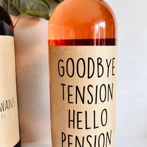 Puede incluir: Una botella de vino de vidrio transparente llena de vino rosado claro. La botella presenta una etiqueta de papel color canela con texto negro que dice: GOODBYE TENSION HELLO PENSION CONGRATS ON RETIRING. Tiene una tapa negra y forma cil&iacute;ndrica.
