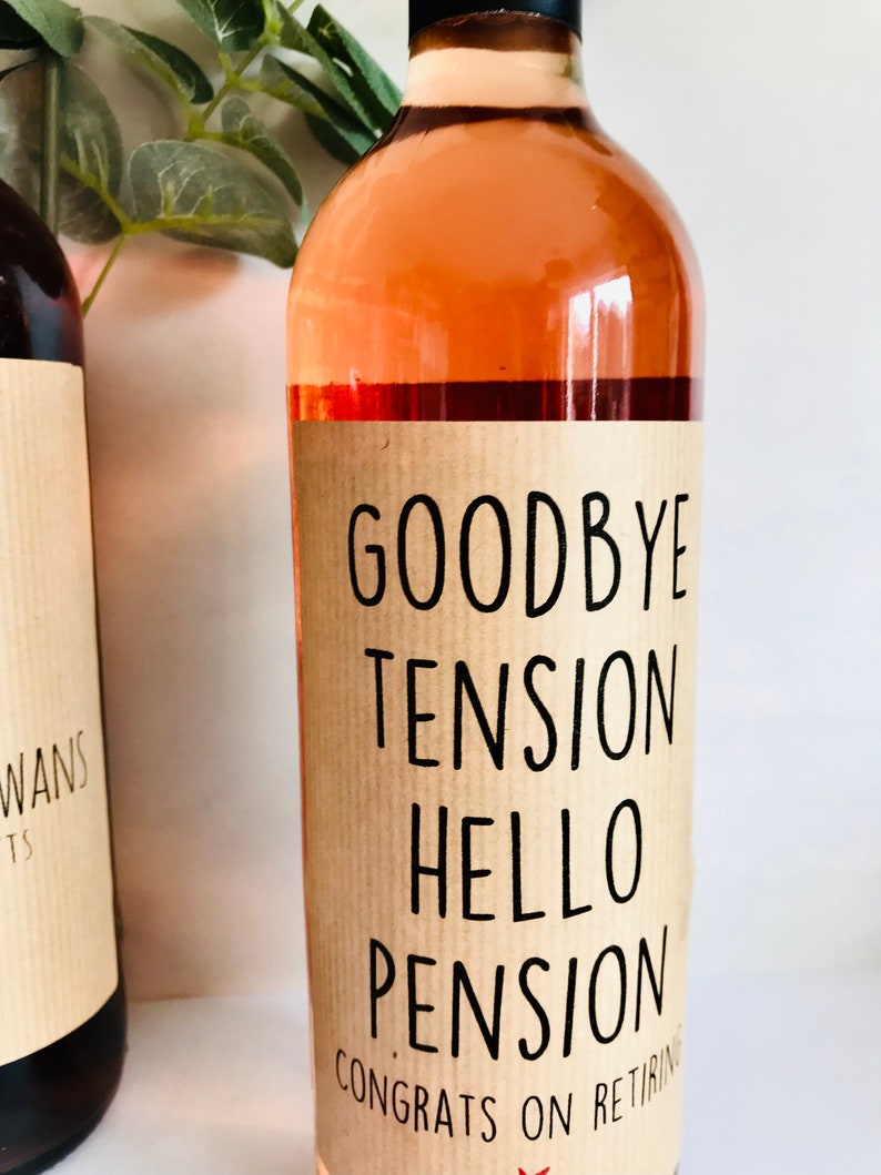 Puede incluir: Una botella de vino de vidrio transparente llena de vino rosado claro. La botella presenta una etiqueta de papel color canela con texto negro que dice: GOODBYE TENSION HELLO PENSION CONGRATS ON RETIRING. Tiene una tapa negra y forma cil&iacute;ndrica.