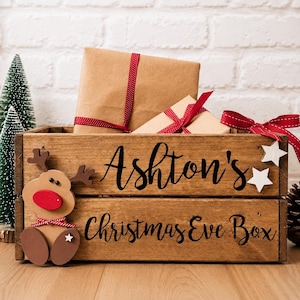 Puede incluir: Una caja de madera rectangular con el texto negro Ashton's Christmas Eve Box impreso en el frente. Un recorte de reno de madera con una nariz roja est&aacute; unido al lado izquierdo, y dos peque&ntilde;as estrellas blancas est&aacute;n unidas a la derecha.