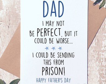 May include: A white card with blue text that reads "DAD I MAY NOT BE PERFECT, BUT IT COULD BE WORSE... I COULD BE SENDING THIS FROM PRISON! HAPPY FATHER'S DAY" with three blue stars above the word "WORSE". The card is on a brown wooden surface with a green plant in the background.