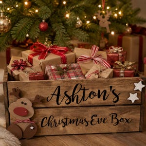 Puede incluir: Una caja de madera con el texto Ashton's Christmas Eve Box, que presenta un recorte de reno y estrellas blancas. La caja est&aacute; llena de varios regalos envueltos, incluyendo cajas con cintas rojas, patrones de cuadros y cordel, bajo un &aacute;rbol.