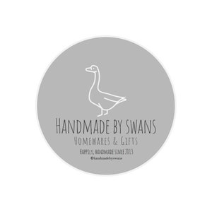 May include: A gray circle with a white outline, featuring a line drawing of a goose. The text "HANDMADE BY SWANS" is written in black below the goose, followed by "HOMEWARES & GIFTS" and "Happily, Handmade Since 2013". The text "@handmadebyswans" is written in black at the bottom of the circle.