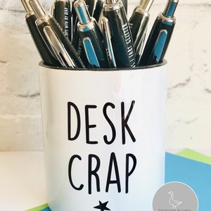 May include: White ceramic desk organiser with black text that reads "DESK CRAP" and a black star. The organiser is filled with black pens.