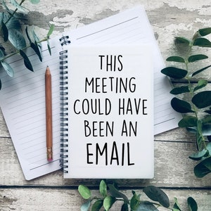 May include: A white notebook with a black spiral binding and the text "THIS MEETING COULD HAVE BEEN AN EMAIL" written on it. The notebook is on a wooden surface with green leaves.