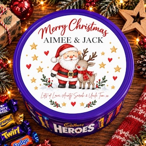 以下が含まれることがあります： 紫色の丸いキャドバリー・ヒーローズの缶。白い蓋には「Merry Christmas, AIMEE & JACK」および「Lots of Love, Aunty Sarah & Uncle Tom」の文字が印刷されています。蓋にはサンタクロースとトナカイのイラストが描かれ、金色の星と赤いハートが散りばめられています。