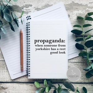 May include: A white notebook with the word "propaganda" and a definition printed on the cover. A wooden pencil and a lined notepad are next to the notebook. Green leaves are in the background.