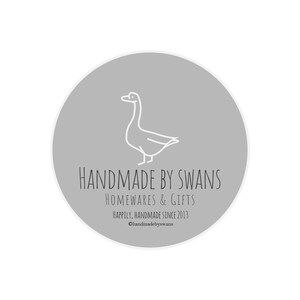 May include: A gray circle with a white outline, featuring a line drawing of a goose. The text "HANDMADE BY SWANS" is written in black below the goose, followed by "HOMEWARES & GIFTS" and "Happily, Handmade Since 2013". The text "@handmadebyswans" is written in black at the bottom of the circle.
