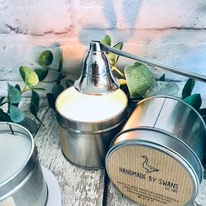 May include: A silver candle snuffer with a long handle is positioned over a silver candle tin. The candle tin has a brown label with the text "HANDMADE BY SWANS EST 2013" and "VANILLA AND WAFFLES WAX CANDLE HAND POURED IN YORKSHIRE www.HANDMADEBYSWANS.COM".
