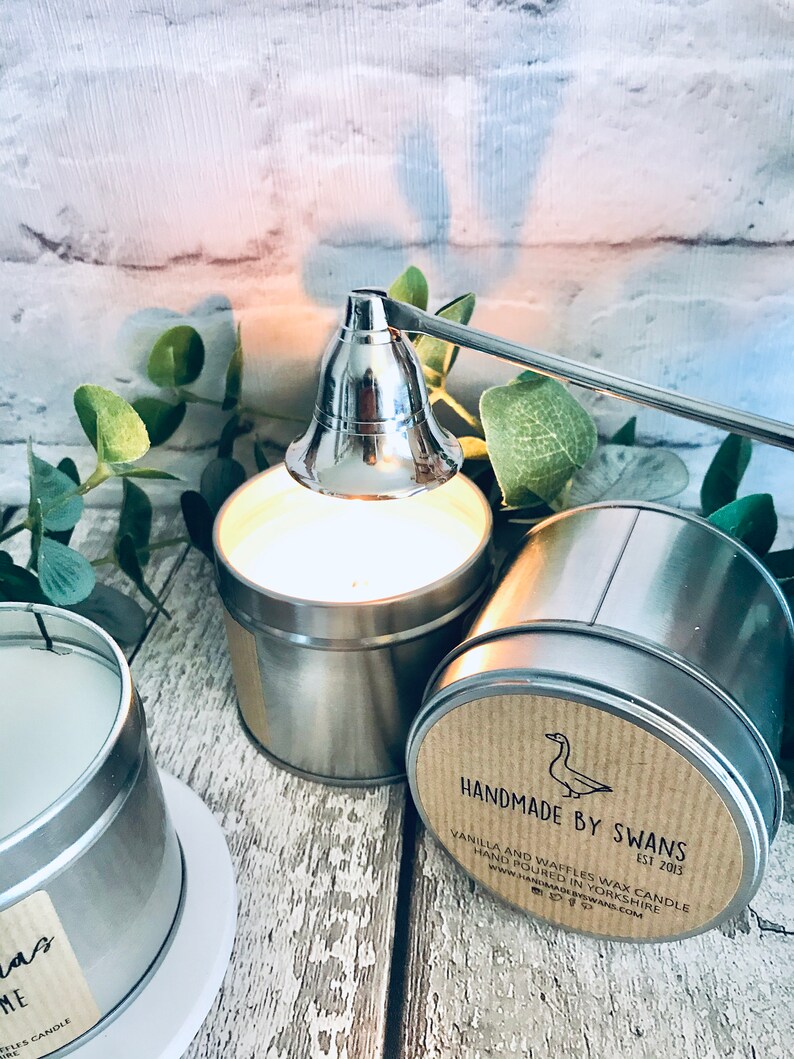 May include: A silver candle snuffer with a long handle is positioned over a silver candle tin. The candle tin has a brown label with the text "HANDMADE BY SWANS EST 2013" and "VANILLA AND WAFFLES WAX CANDLE HAND POURED IN YORKSHIRE www.HANDMADEBYSWANS.COM".
