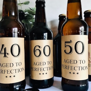 May include: Three brown glass beer bottles with brown paper labels. The labels have black text that reads "40 Aged to Perfection", "60 Aged to Perfection", and "50 Aged to Perfection".
