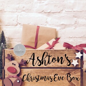 Puede incluir: Una caja de madera rectangular con el texto "Ashton's Christmas Eve Box" en letra cursiva negra. La caja presenta un recorte de reno de madera en el lado izquierdo y dos peque&ntilde;as estrellas de madera blanca en el lado derecho, llena de paquetes de papel marr&oacute;n.