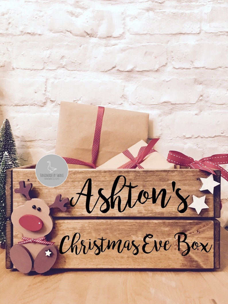 Puede incluir: Una caja de madera rectangular con el texto "Ashton's Christmas Eve Box" en letra cursiva negra. La caja presenta un recorte de reno de madera en el lado izquierdo y dos peque&ntilde;as estrellas de madera blanca en el lado derecho, llena de paquetes de papel marr&oacute;n.