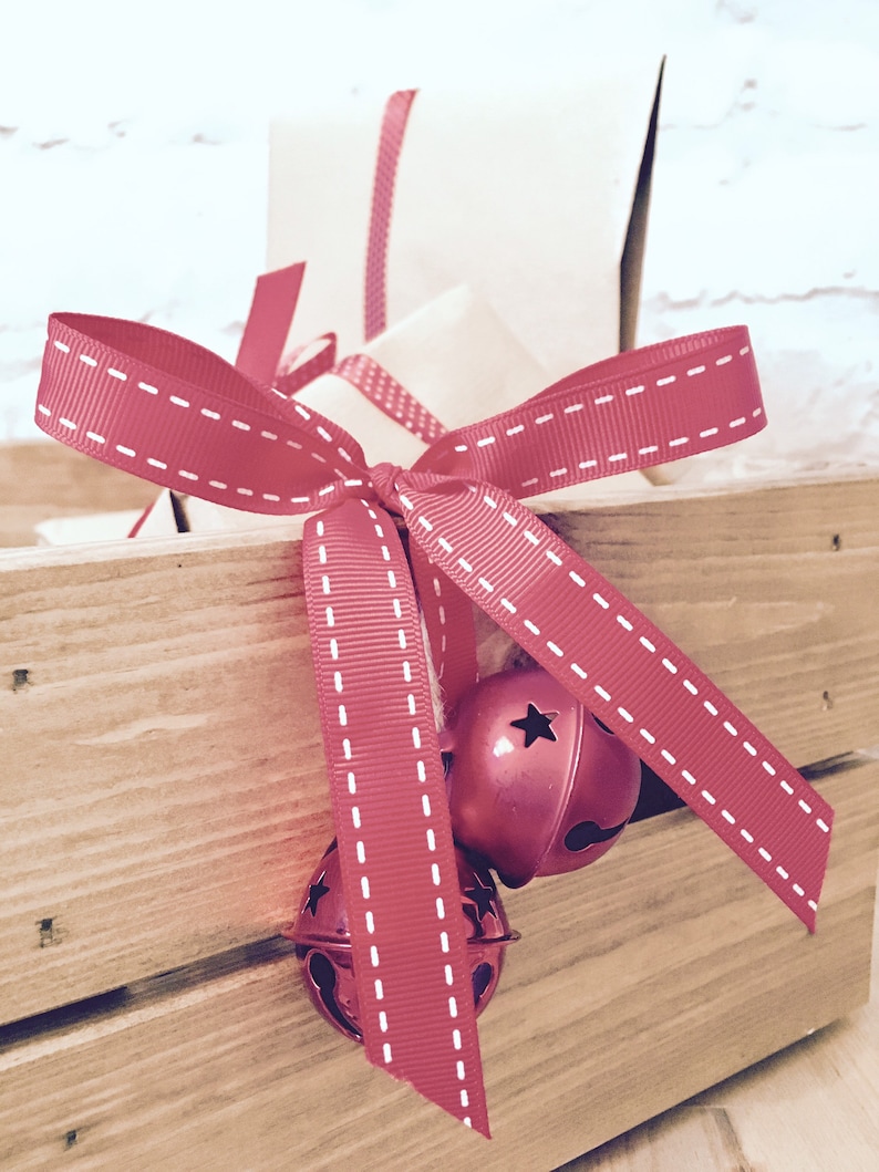 Puede incluir: Una caja de madera de color marr&oacute;n claro que contiene un regalo envuelto en papel beige liso. Una cinta de grosgrain roja con bordes cosidos en blanco est&aacute; atada en un lazo alrededor del paquete, con dos cascabeles rojos met&aacute;licos.
