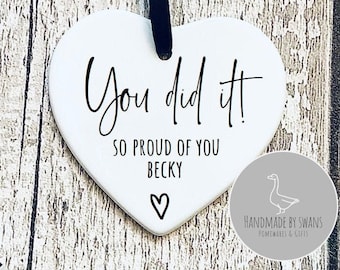 May include: White ceramic heart-shaped ornament with black text that reads "You did it! So proud of you Becky" with a small heart symbol below the text. The ornament is hanging from a black ribbon.