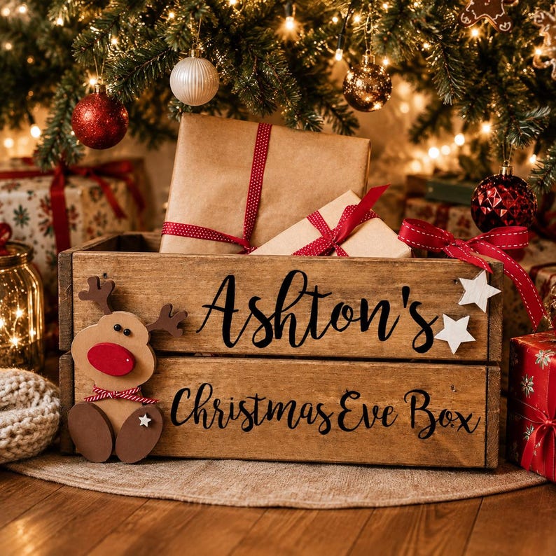 Puede incluir: Una caja de madera rectangular con acabado marr&oacute;n oscuro, que presenta el texto negro "Ashton's Christmas Eve Box" en el frente. Un recorte de reno de madera con nariz roja est&aacute; fijado en el lado izquierdo, y dos peque&ntilde;as estrellas de madera blancas est&aacute;n a la derecha.