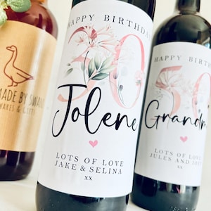 May include: Two wine bottles with personalised labels. The labels are white with pink floral designs and black text. The text on the left label reads "Happy Birthday Jolene Lots of Love Jake & Selina XX". The text on the right label reads "Happy Birthday Grandma Lots of Love Jules and Joe XX".