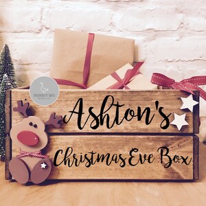 Puede incluir: Una caja de madera rectangular con acabado marr&oacute;n oscuro que presenta el texto negro Ashton's Christmas Eve Box. El frente est&aacute; decorado con una figura de reno de madera con nariz roja y dos peque&ntilde;as estrellas blancas en el lado derecho.